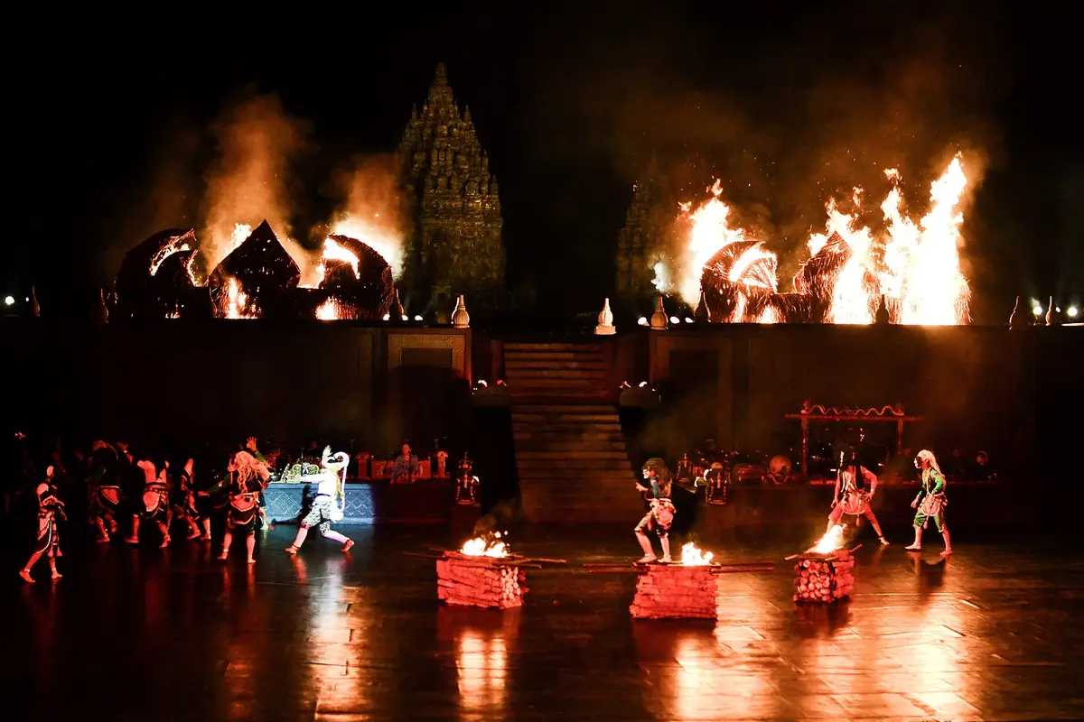 Dancers on the open-air stage at night with fires burning on stage and behind it, and the main Prambanan temple silhouetted in the smoke